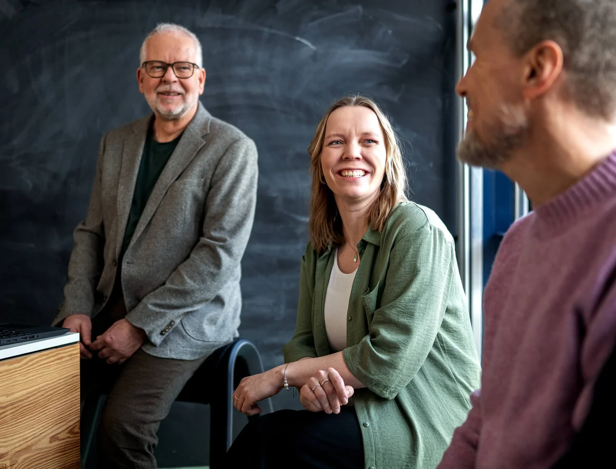 Drei Personen sitzen zusammen in einem Raum und lächeln sich an. Eine Person sitzt links, die andere rechts, und die dritte Person zwischen ihnen. Die Atmosphäre wirkt entspannt und freundlich. Im Hintergrund ist eine Tafel zu sehen.