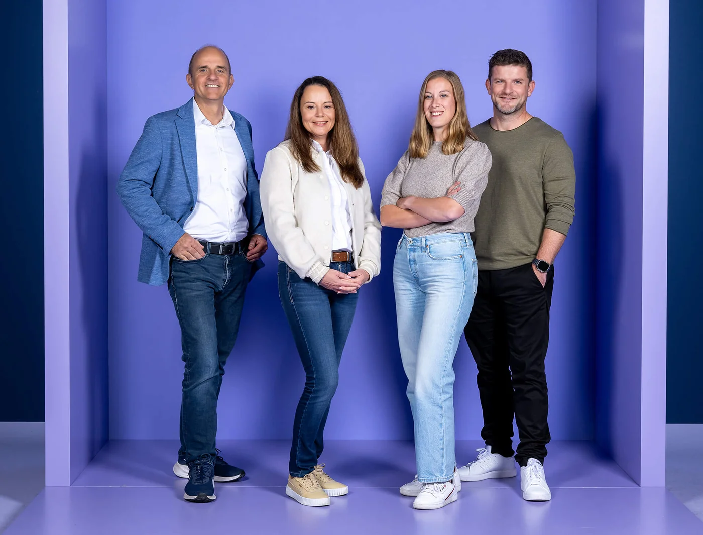 Vier Personen stehen vor einem lila Hintergrund. Sie wirken entspannt und freundlich. Die Gruppe besteht aus zwei Männern und zwei Frauen, die lässige Kleidung tragen.