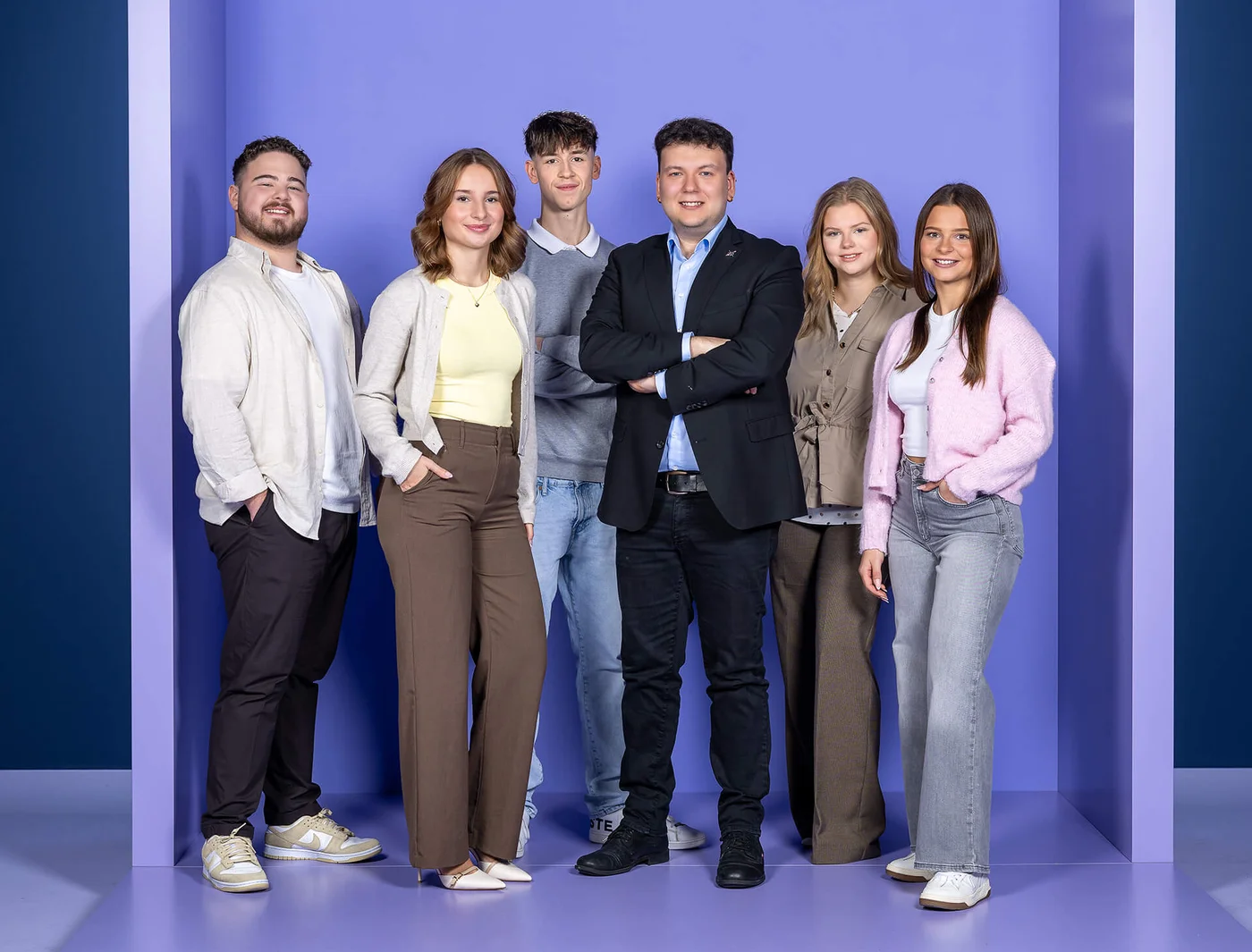 Eine Gruppe von sechs jungen Erwachsenen steht vor einem lila Hintergrund und lächelt in die Kamera. Drei Frauen und drei Männer tragen legere Kleidung. Der Mann in der Mitte trägt einen Anzug und hält die Arme verschränkt. 