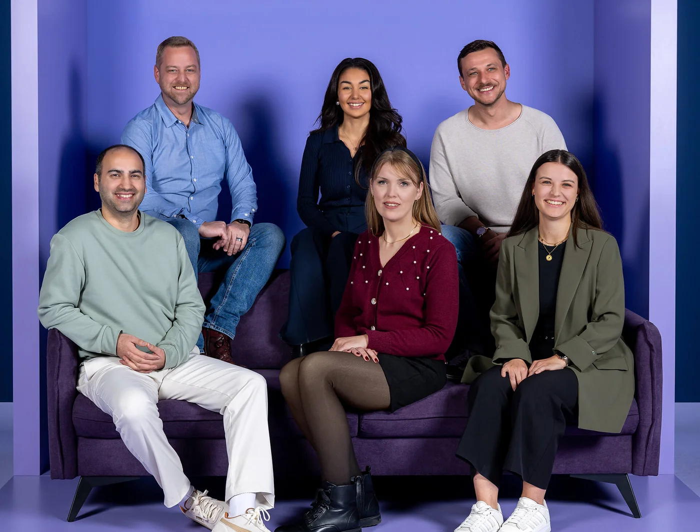 Entschuldigung, ich weiß nicht, wer auf dem Foto ist. Einfacher Hintergrund mit sechs lächelnden Menschen, die in zwei Reihen auf einem lila Sofa sitzen. Drei stehen dahinter und drei sitzen davor. Alle sind lässig gekleidet.