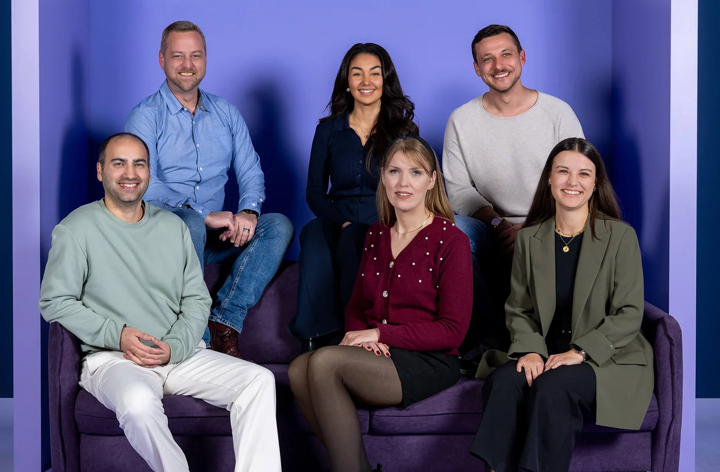 Entschuldigung, ich weiß nicht, wer auf dem Foto ist. Einfacher Hintergrund mit sechs lächelnden Menschen, die in zwei Reihen auf einem lila Sofa sitzen. Drei stehen dahinter und drei sitzen davor. Alle sind lässig gekleidet.