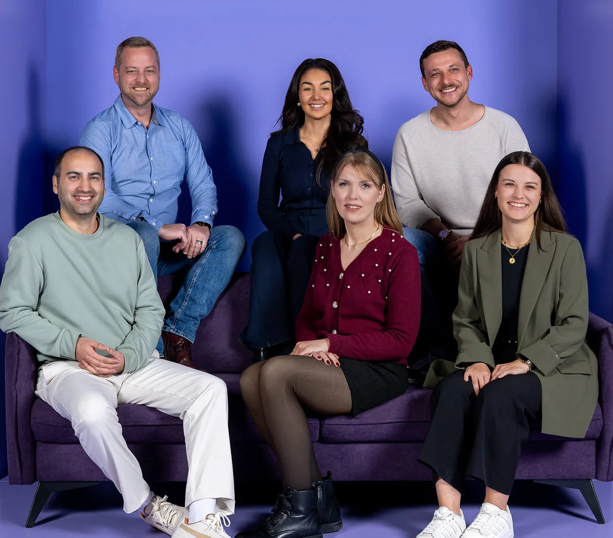 Entschuldigung, ich weiß nicht, wer auf dem Foto ist. Einfacher Hintergrund mit sechs lächelnden Menschen, die in zwei Reihen auf einem lila Sofa sitzen. Drei stehen dahinter und drei sitzen davor. Alle sind lässig gekleidet.