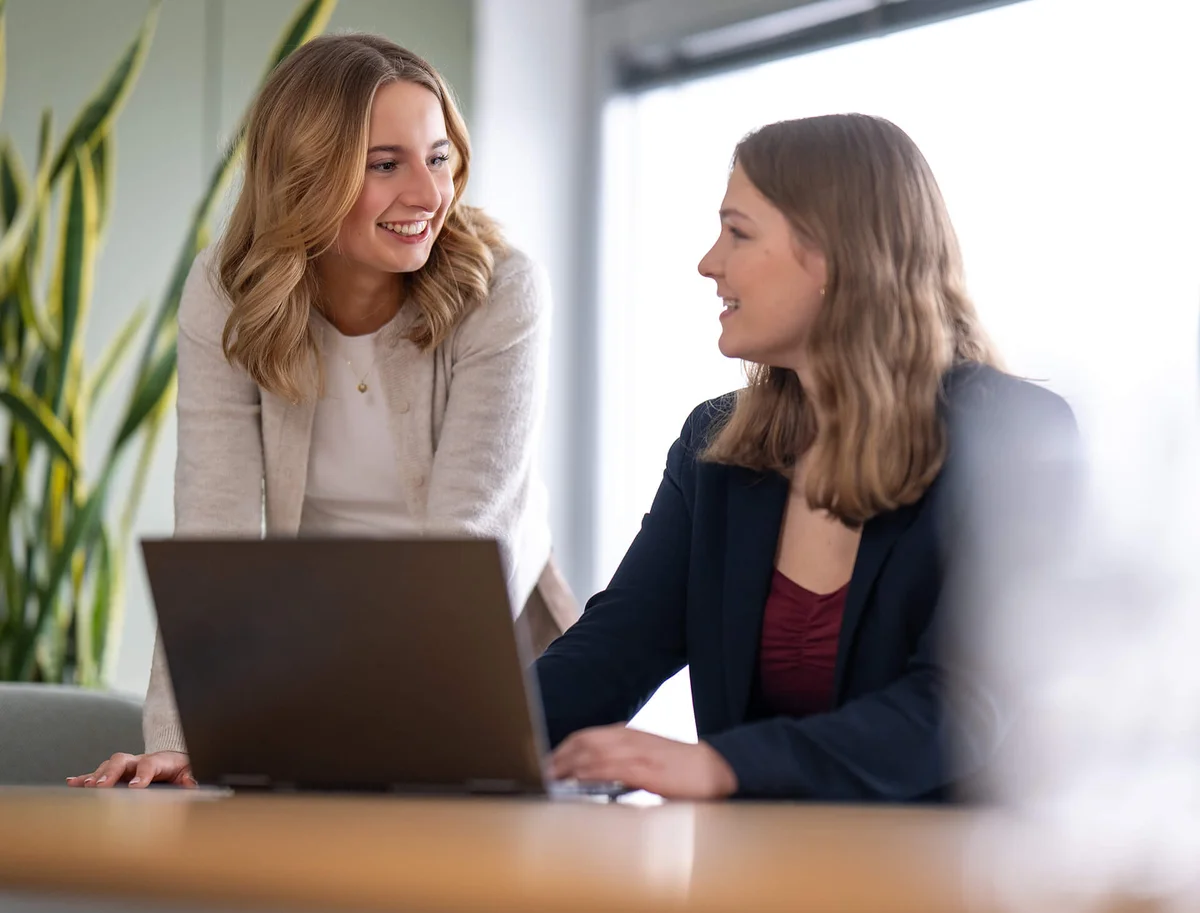 Zwei Frauen in einem Büro, eine steht und lächelt, während sie mit der anderen spricht, die an einem Laptop arbeitet. Beide scheinen sich in einem freundlichen Gespräch zu befinden.