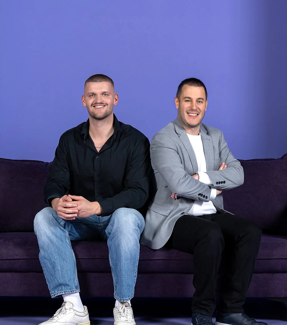 Zwei Männer sitzen auf einem lila Sofa vor lila Hintergrund. Der Mann links trägt ein schwarzes Hemd und Jeans, der rechts einen grauen Blazer und schwarze Hose. Beide lächeln.