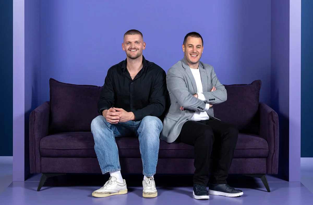 Zwei Männer sitzen auf einem lila Sofa vor lila Hintergrund. Der Mann links trägt ein schwarzes Hemd und Jeans, der rechts einen grauen Blazer und schwarze Hose. Beide lächeln.
