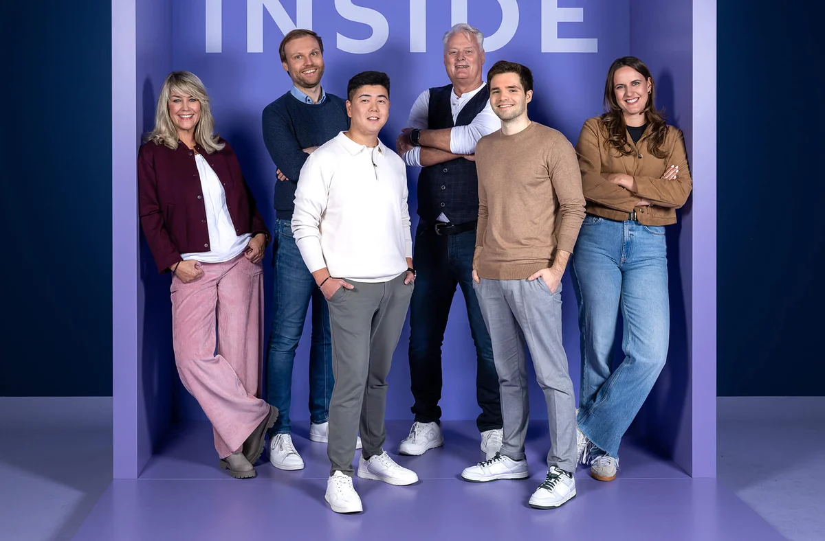 Eine Gruppe von fünf Menschen steht entspannt lächelnd in einem lilafarbenen Raum mit der Aufschrift "INSIDE" an der Wand. Sie tragen lässige, modische Kleidung und unterschiedliche Posen. Die Stimmung wirkt freundlich und einladend.