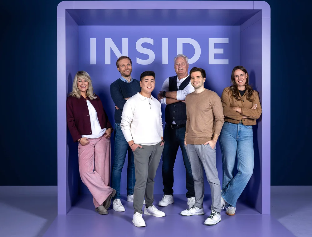 Eine Gruppe von fünf Menschen steht entspannt lächelnd in einem lilafarbenen Raum mit der Aufschrift "INSIDE" an der Wand. Sie tragen lässige, modische Kleidung und unterschiedliche Posen. Die Stimmung wirkt freundlich und einladend.