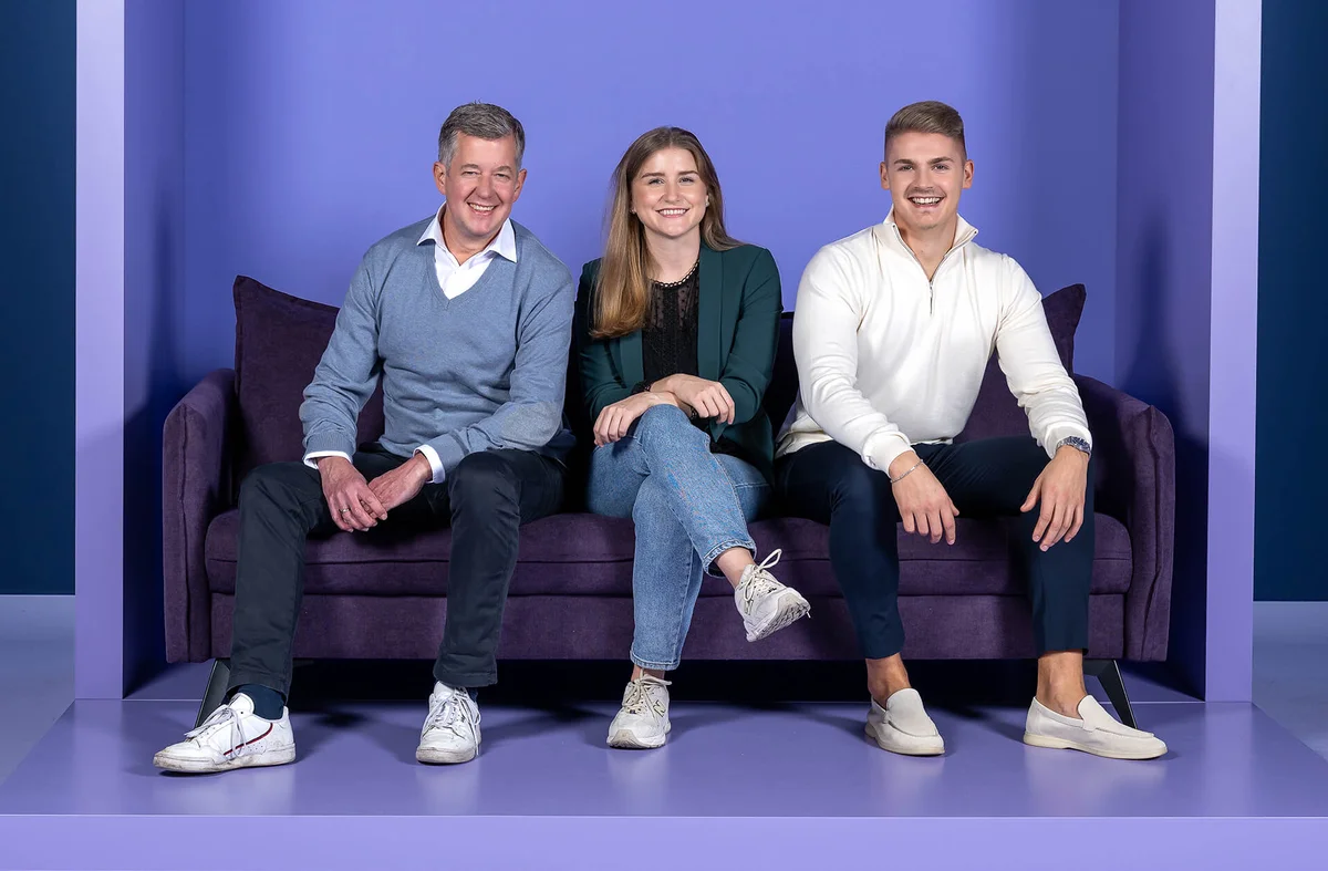 Eine Frau und zwei Männer sitzen lächelnd auf einem lila Sofa. Sie sind lässig gekleidet, der Hintergrund ist ebenfalls lila.