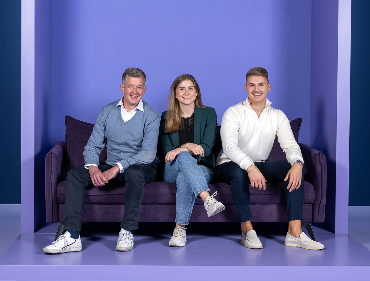 Eine Frau und zwei Männer sitzen lächelnd auf einem lila Sofa. Sie sind lässig gekleidet, der Hintergrund ist ebenfalls lila.