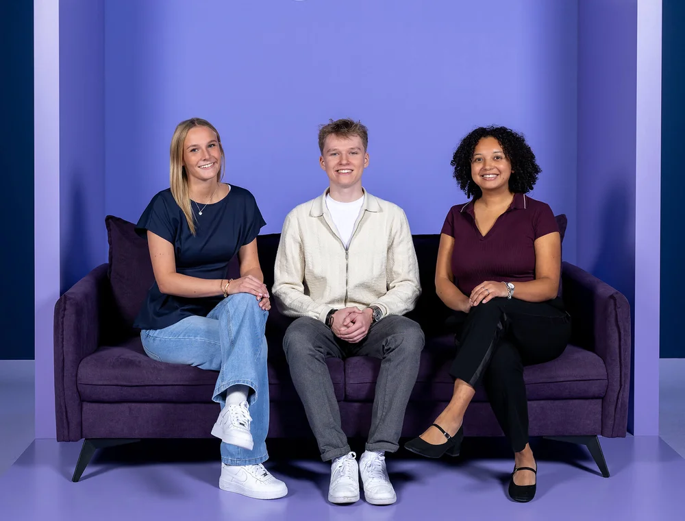 Drei Personen sitzen lächelnd auf einem lila Sofa vor einem lila Hintergrund. Von links nach rechts: eine Frau in Jeans und blauem Oberteil, ein Mann in heller Jacke, und eine Frau in schwarzer Hose und dunkelrotem Oberteil. Alle wirken entspannt und freundlich.