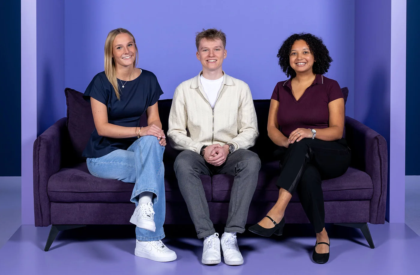 Drei Personen sitzen lächelnd auf einem lila Sofa vor einem lila Hintergrund. Von links nach rechts: eine Frau in Jeans und blauem Oberteil, ein Mann in heller Jacke, und eine Frau in schwarzer Hose und dunkelrotem Oberteil. Alle wirken entspannt und freundlich.