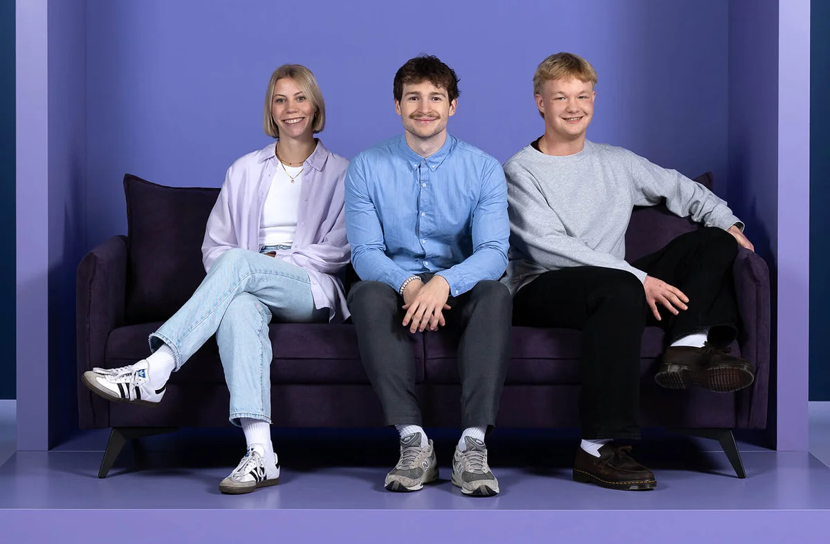 Drei Personen sitzen auf einem dunkellila Sofa vor einem lila Hintergrund mit dem Wort "INSIDE" darüber. Die mittlere Person trägt ein hellblaues Hemd, die linke eine weiße Jacke, und die rechte einen grauen Pullover. Alle lächeln und wirken entspannt.