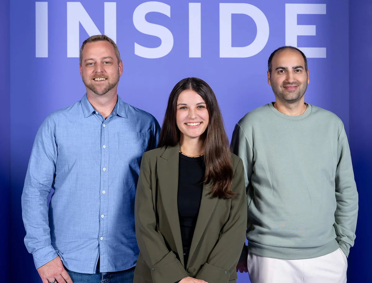 Drei lächelnde Personen stehen vor einem lila Hintergrund mit dem Wort "INSIDE" oben sichtbar. Die mittlere Person ist eine Frau in einem dunkelgrünen Blazer, die anderen beiden sind Männer in einem blauen Hemd und einem hellgrünen Pullover. Sie wirken freundlich und entspannt.