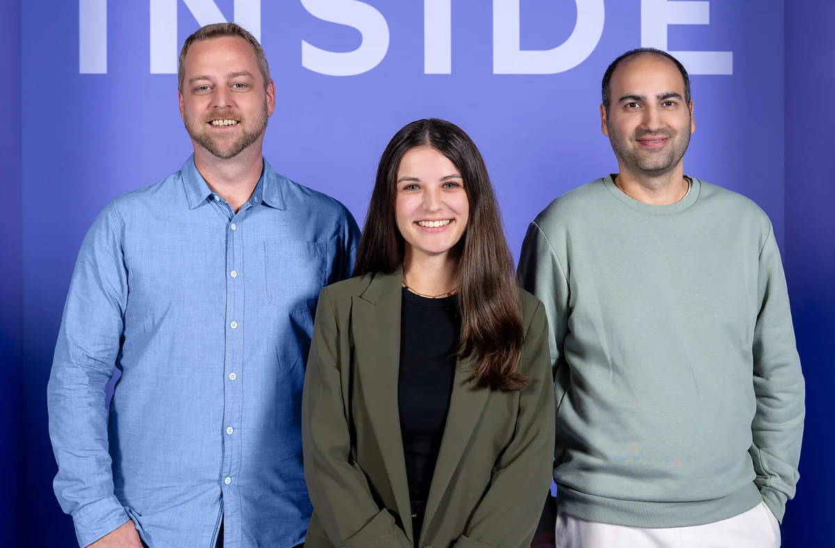 Drei lächelnde Personen stehen vor einem lila Hintergrund mit dem Wort "INSIDE" oben sichtbar. Die mittlere Person ist eine Frau in einem dunkelgrünen Blazer, die anderen beiden sind Männer in einem blauen Hemd und einem hellgrünen Pullover. Sie wirken freundlich und entspannt.