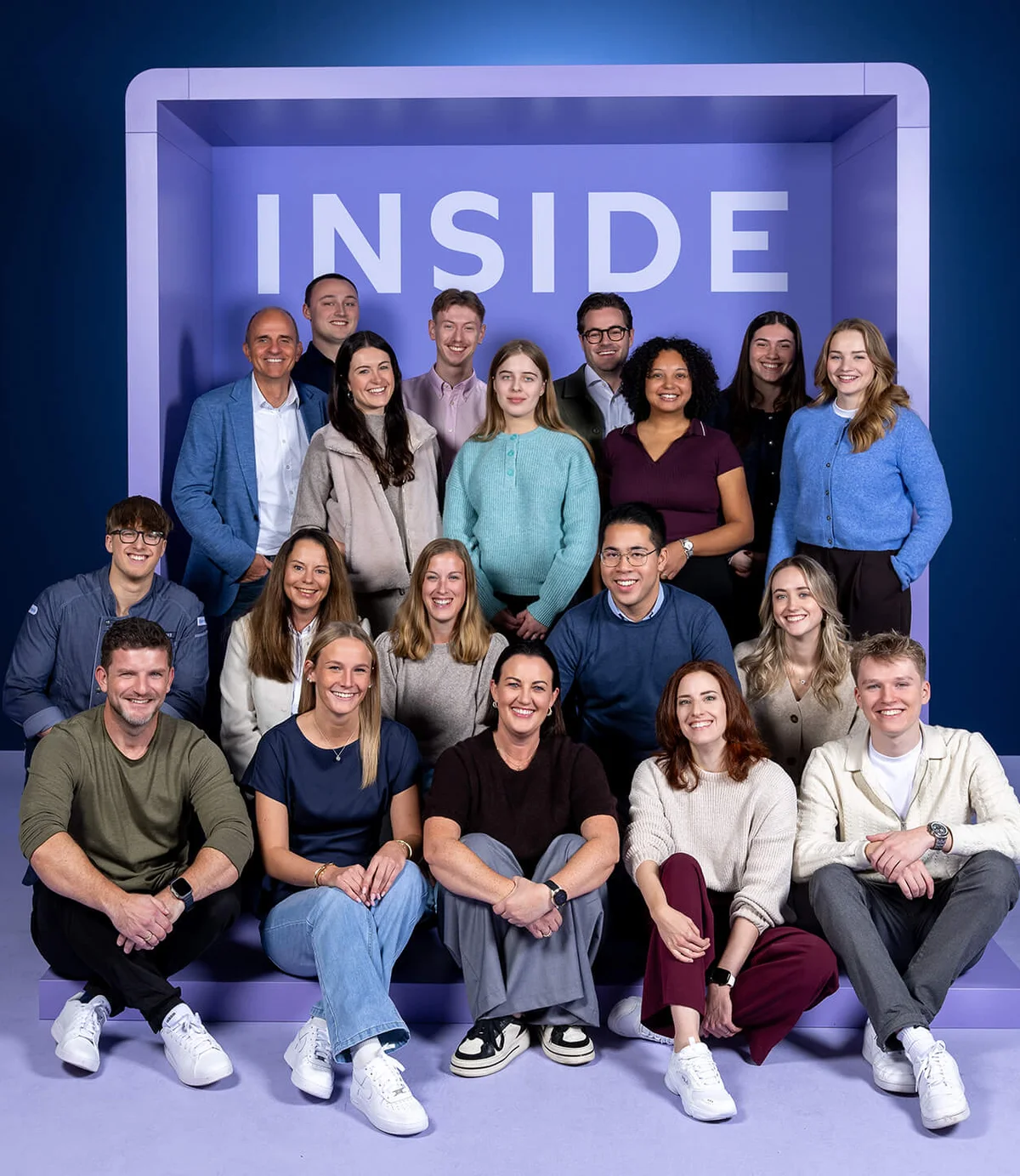 Eine Gruppe von 19 lächelnden Menschen posiert vor einem blauen Hintergrund in einem großen, offenen Rahmen mit der Aufschrift "INSIDE". Die Personen sind in zwei Reihen, sitzend und stehend, angeordnet.
