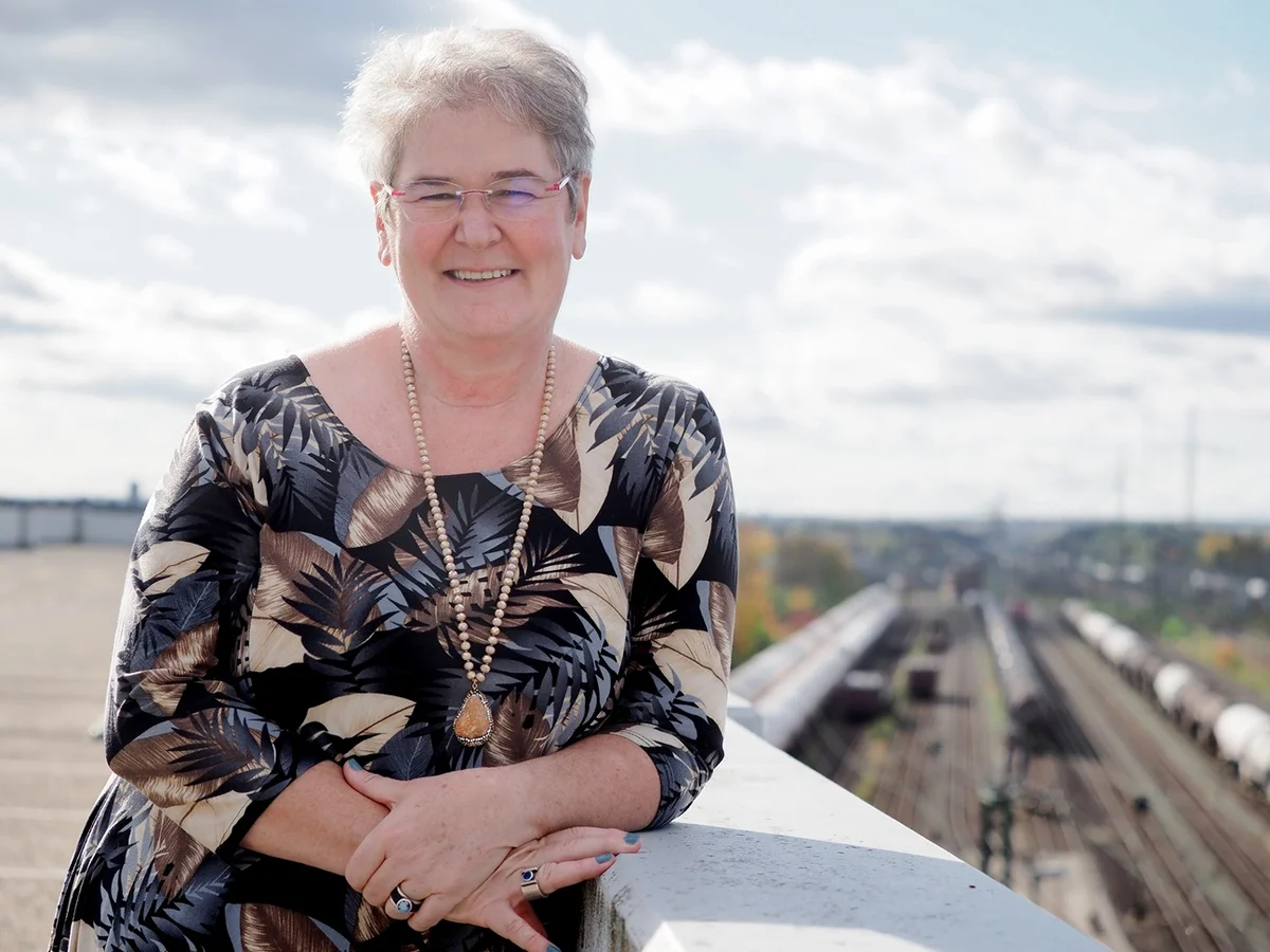Eine lächelnde ältere Frau steht an einem Geländer im Freien. Sie trägt ein gemustertes Oberteil und eine Halskette. Im Hintergrund sieht man Eisenbahnschienen und einen bewölkten Himmel.