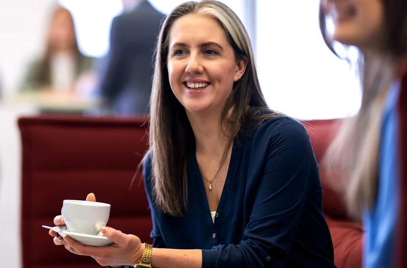 Eine Frau sitzt lächelnd auf einem roten Sofa und hält eine Tasse in der Hand. Im Hintergrund sind unscharf andere Menschen zu sehen, die im Raum stehen. Die Atmosphäre wirkt entspannt und freundlich.
