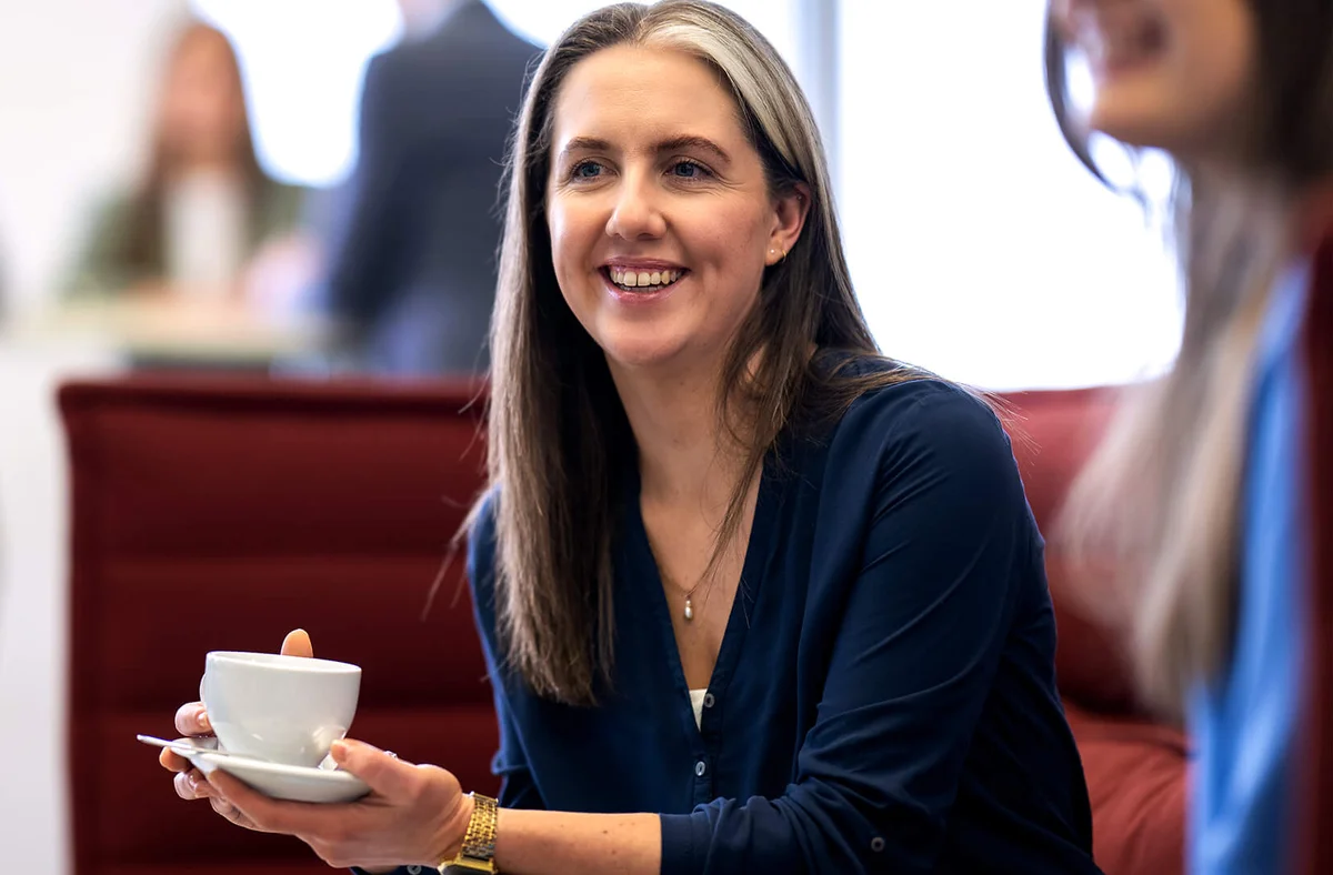 Eine Frau sitzt lächelnd auf einem roten Sofa und hält eine Tasse in der Hand. Im Hintergrund sind unscharf andere Menschen zu sehen, die im Raum stehen. Die Atmosphäre wirkt entspannt und freundlich.