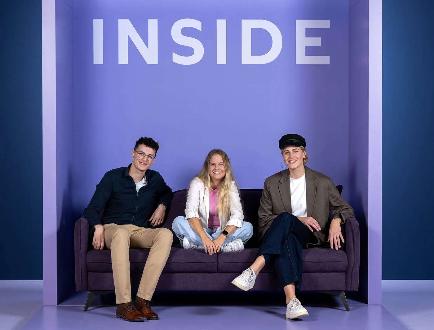 Drei Personen sitzen auf einem lila Sofa vor einem lila Hintergrund mit dem großen Wort "INSIDE" darüber. Die Personen lächeln und sind lässig gekleidet. Die Stimmung wirkt entspannt und freundlich.