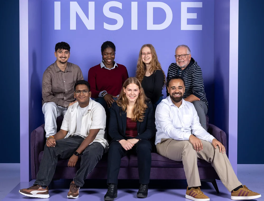 Sieben Personen sitzen und stehen vor einem lila Hintergrund mit dem Wort "INSIDE". Sie lächeln und tragen verschiedene legere und formelle Kleidung.