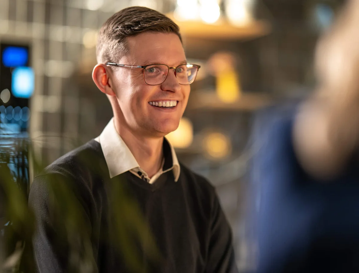 Es tut mir leid, ich weiß nicht, wer die Person auf dem Bild ist. Ein Mann mit Brille und Hemd, der lächelt. Der Hintergrund ist unscharf und zeigt möglicherweise ein beleuchtetes Regal.