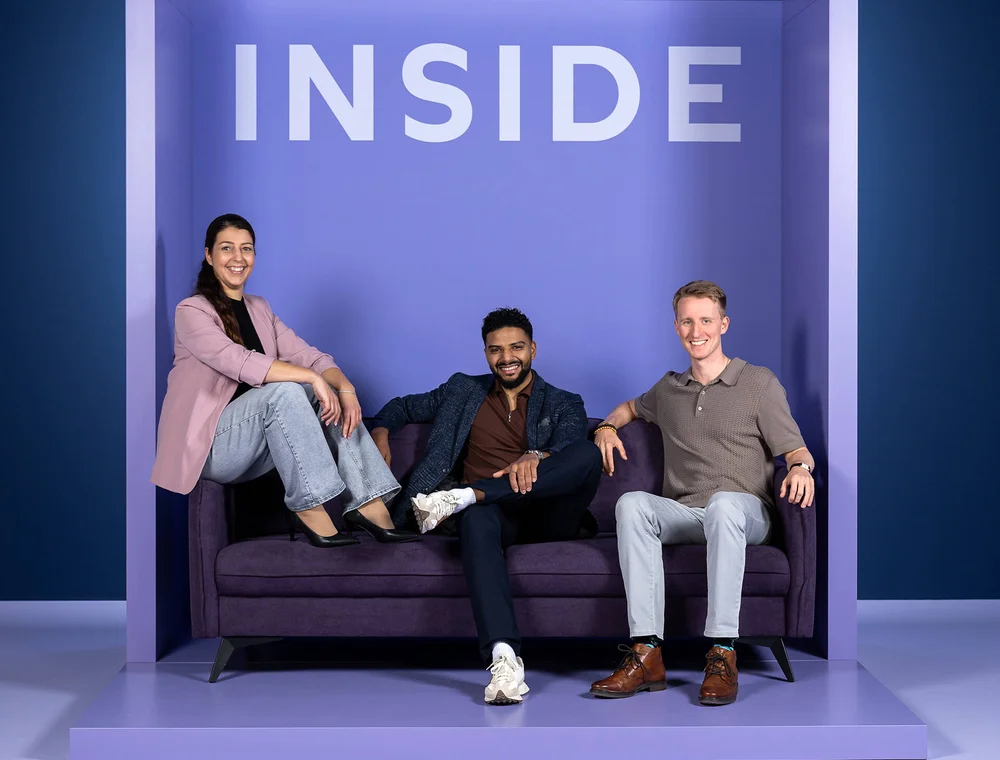 Eine Frau und zwei Männer sitzen lächelnd auf einem lila Sofa. Hinter ihnen steht das Wort "INSIDE" in großen Buchstaben. Alle wirken entspannt und freundlich.