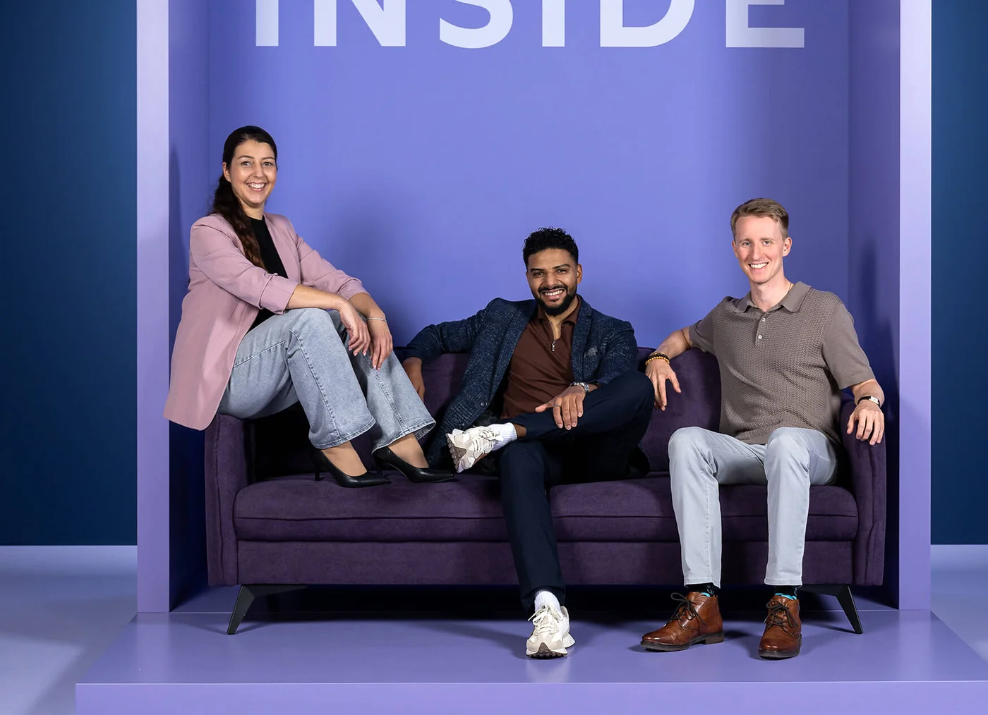 Eine Frau und zwei Männer sitzen lächelnd auf einem lila Sofa. Hinter ihnen steht das Wort "INSIDE" in großen Buchstaben. Alle wirken entspannt und freundlich.