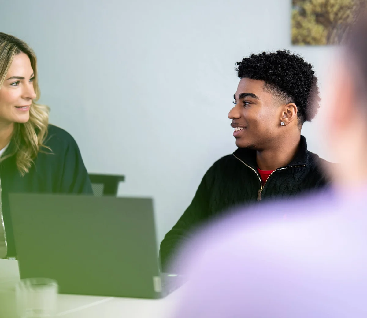 Zwei Personen sitzen an einem Tisch mit einem Laptop und unterhalten sich lächelnd miteinander. Die Atmosphäre wirkt entspannt und freundlich.