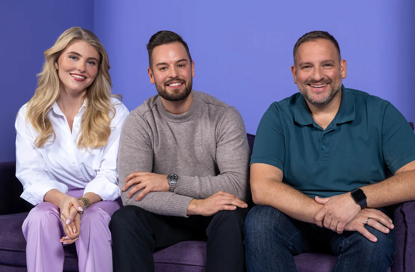 Drei Personen sitzen auf einem lila Sofa vor einem blauen Hintergrund. Zwei Männer und eine Frau lächeln in die Kamera. Die Frau trägt ein weißes Hemd und helle Hosen. Einer der Männer trägt einen grauen Pullover, der andere ein blaues Poloshirt.