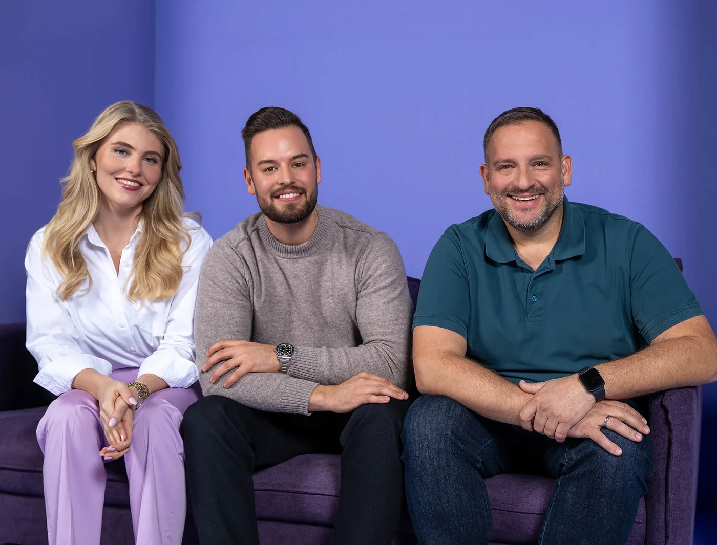 Drei Personen sitzen auf einem lila Sofa vor einem blauen Hintergrund. Zwei Männer und eine Frau lächeln in die Kamera. Die Frau trägt ein weißes Hemd und helle Hosen. Einer der Männer trägt einen grauen Pullover, der andere ein blaues Poloshirt.