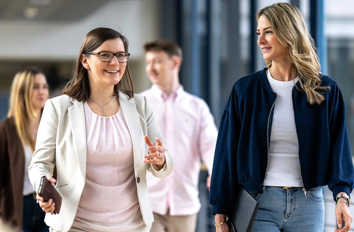Zwei Frauen unterhalten sich lächelnd, während sie einen Büroflur entlanggehen. Beide tragen geschäftliche Kleidung. Im Hintergrund sind zwei weitere Personen zu sehen, die ebenfalls lächeln. Die Atmosphäre wirkt freundlich und professionell.