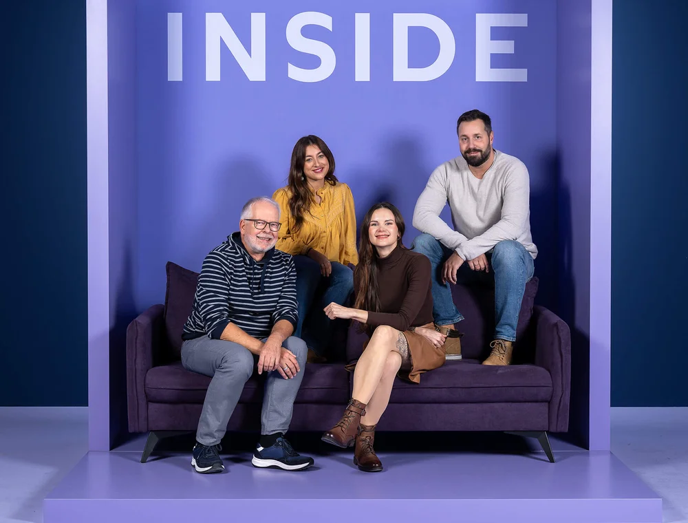 Vier Personen sitzen und stehen auf einem lila Sofa vor einer Wand mit dem Schriftzug „INSIDE“. Drei Menschen sitzen auf dem Sofa, während einer auf der rechten Sofalehne sitzt. Alle sind entspannt und lächeln.