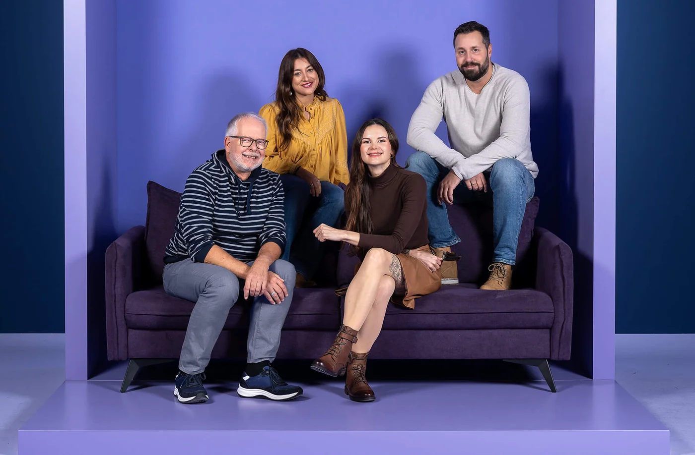 Vier Personen sitzen und stehen auf einem lila Sofa vor einer Wand mit dem Schriftzug „INSIDE“. Drei Menschen sitzen auf dem Sofa, während einer auf der rechten Sofalehne sitzt. Alle sind entspannt und lächeln.