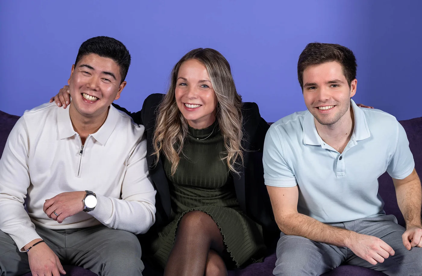 Drei Personen sitzen nebeneinander vor einem blauen Hintergrund und lächeln in die Kamera. Die Person in der Mitte hat die Arme um die beiden anderen gelegt. Alle wirken fröhlich und entspannt.