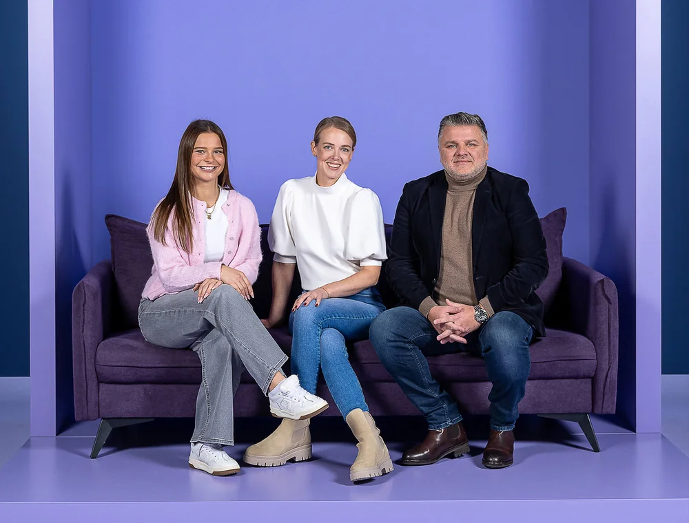 Drei Personen sitzen auf einem lila Sofa vor einem lila Hintergrund. Zwei Frauen und ein Mann tragen lässige Kleidung. Sie lächeln und blicken direkt in die Kamera.
