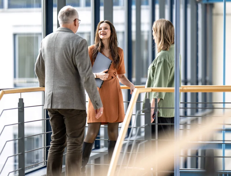 Drei Personen stehen in einer modernen Büro-Umgebung und unterhalten sich freundlich. Eine Frau in der Mitte hält einen Laptop und lächelt. Die anderen beiden Personen hören zu. Alle stehen auf einem erhöhten Gang mit einem Geländer.