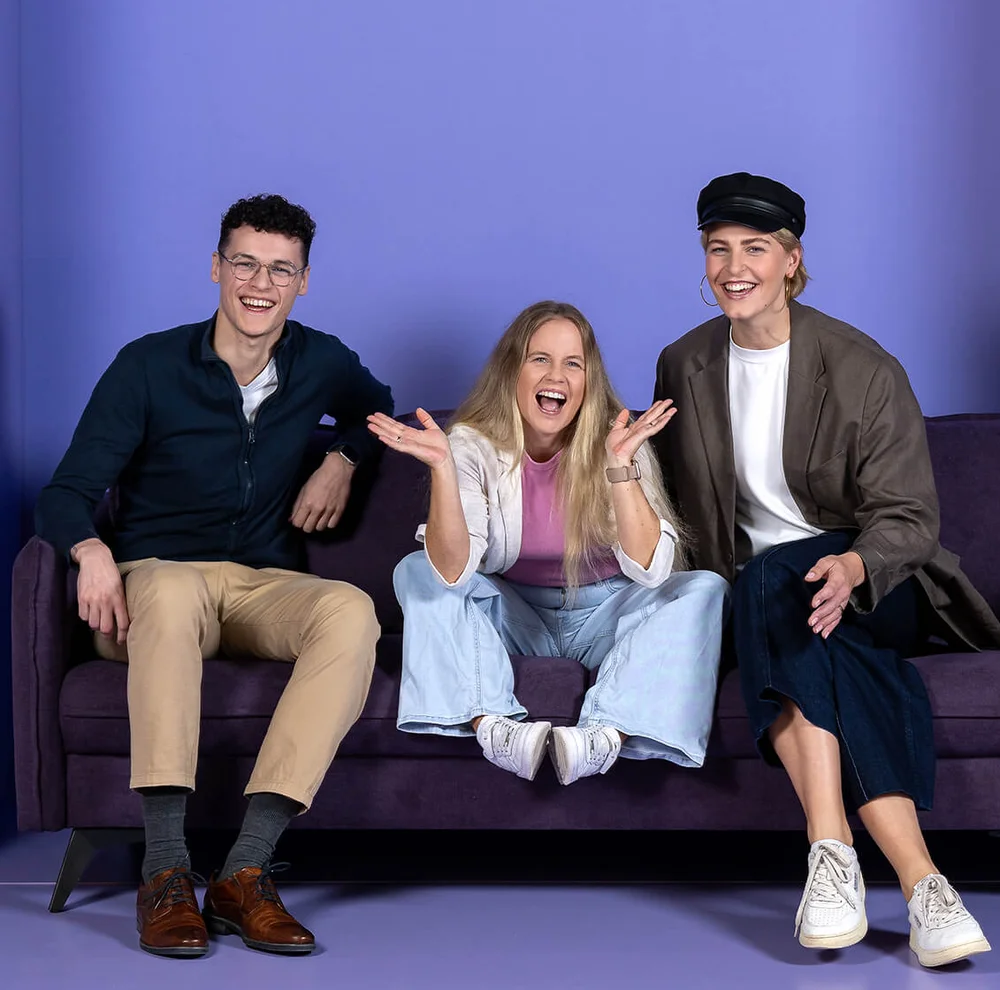 Drei fröhliche Personen sitzen auf einem lila Sofa unter dem Schriftzug "INSIDE". Die mittlere Person gestikuliert enthusiastisch, während die anderen zwei lächeln. Der Hintergrund ist in Lila- und Blautönen gehalten.