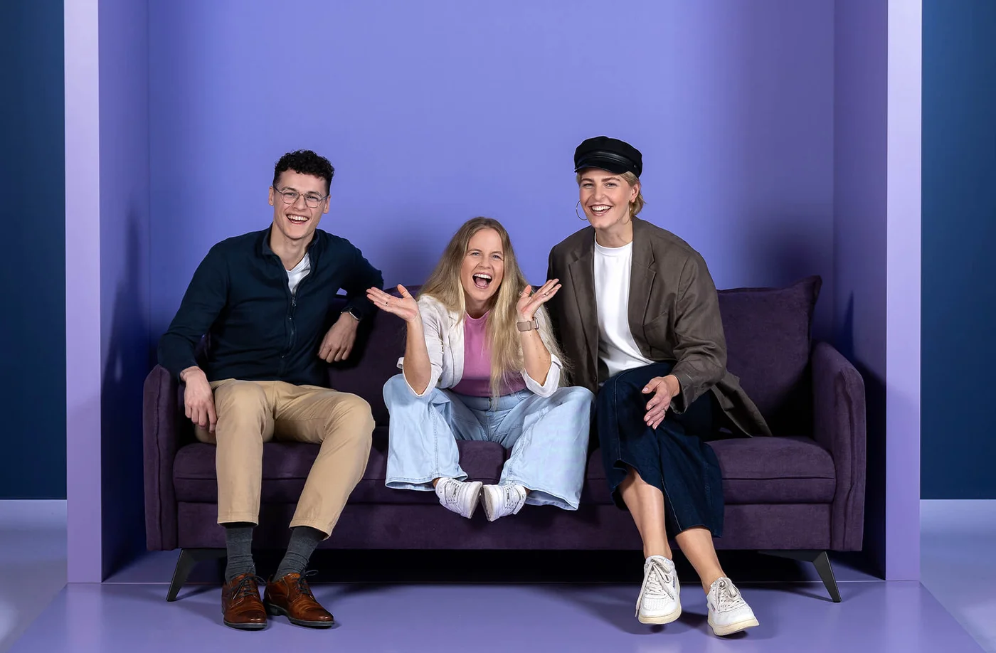 Drei fröhliche Personen sitzen auf einem lila Sofa unter dem Schriftzug "INSIDE". Die mittlere Person gestikuliert enthusiastisch, während die anderen zwei lächeln. Der Hintergrund ist in Lila- und Blautönen gehalten.