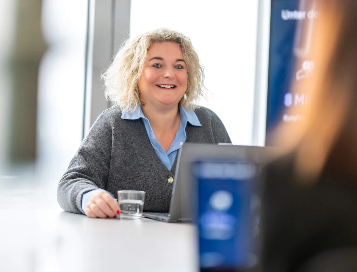 Eine lächelnde Frau mit blondem lockigem Haar sitzt an einem Tisch in einem Büro. Vor ihr stehen ein Laptop und ein Glas Wasser. Sie schaut eine andere Person an, die teilweise sichtbar ist. Die Atmosphäre wirkt freundlich und professionell.