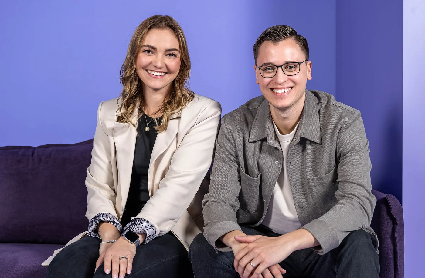 Zwei Personen sitzen nebeneinander auf einem violetten Sofa vor einem blauen Hintergrund. Beide lächeln in die Kamera. Die Frau trägt einen cremefarbenen Blazer, der Mann eine graue Jacke.