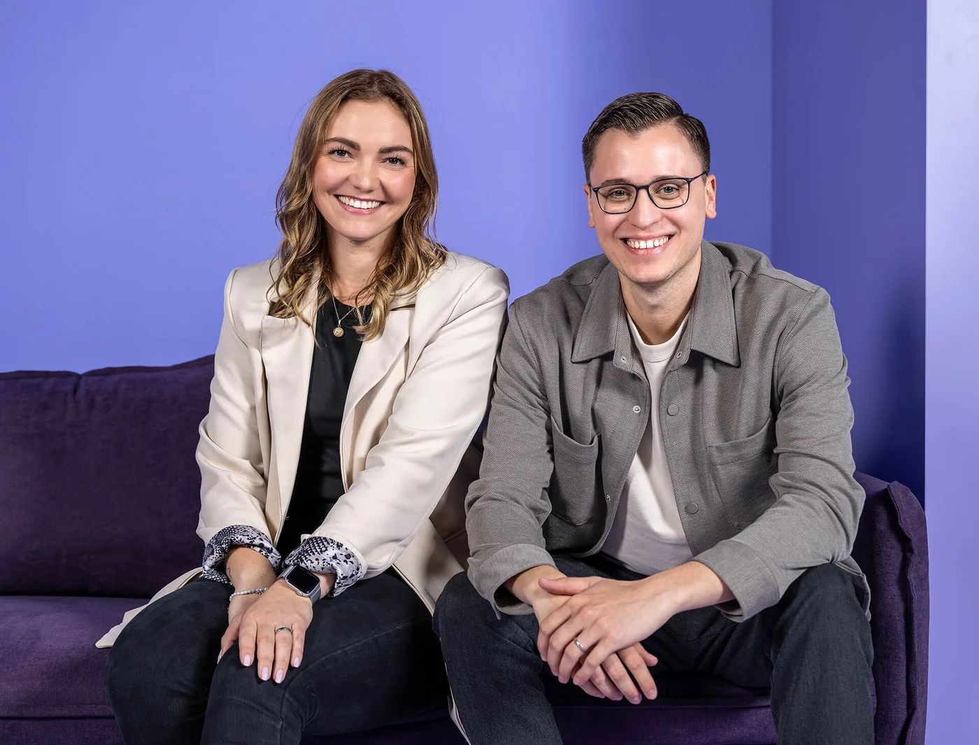 Zwei Personen sitzen nebeneinander auf einem violetten Sofa vor einem blauen Hintergrund. Beide lächeln in die Kamera. Die Frau trägt einen cremefarbenen Blazer, der Mann eine graue Jacke.