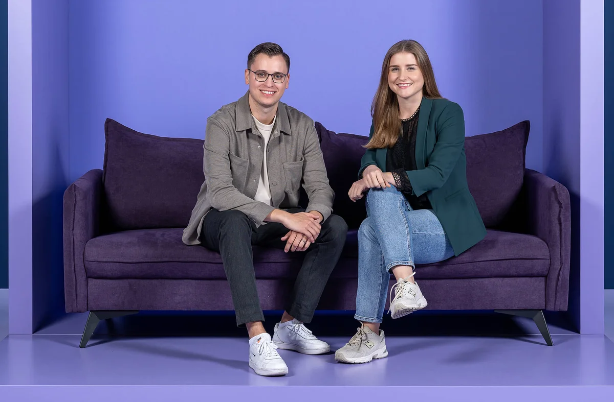 Ein Mann und eine Frau sitzen auf einem lila Sofa vor einem einfarbigen Hintergrund. Beide lächeln in die Kamera. Der Mann trägt eine Brille und Freizeitkleidung, die Frau hat langes Haar und trägt einen Blazer und Jeans.
