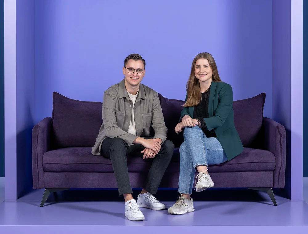 Ein Mann und eine Frau sitzen auf einem lila Sofa vor einem einfarbigen Hintergrund. Beide lächeln in die Kamera. Der Mann trägt eine Brille und Freizeitkleidung, die Frau hat langes Haar und trägt einen Blazer und Jeans.