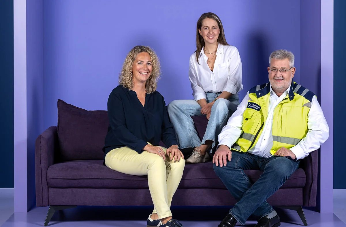 Drei Personen sitzen auf einem lila Sofa vor einem lila Hintergrund. Zwei Frauen tragen legere Kleidung, eine sitzt auf der Lehne. Ein Mann in einer gelben Sicherheitsweste sitzt rechts. Alle lächeln.