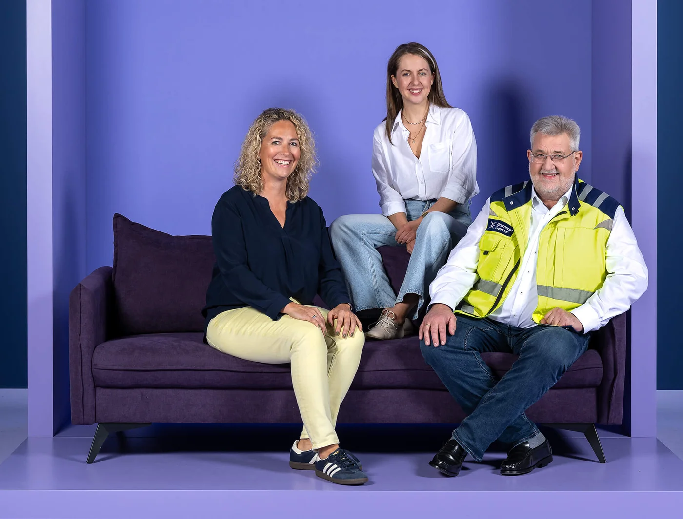 Drei Personen sitzen auf einem lila Sofa vor einem lila Hintergrund. Zwei Frauen tragen legere Kleidung, eine sitzt auf der Lehne. Ein Mann in einer gelben Sicherheitsweste sitzt rechts. Alle lächeln.