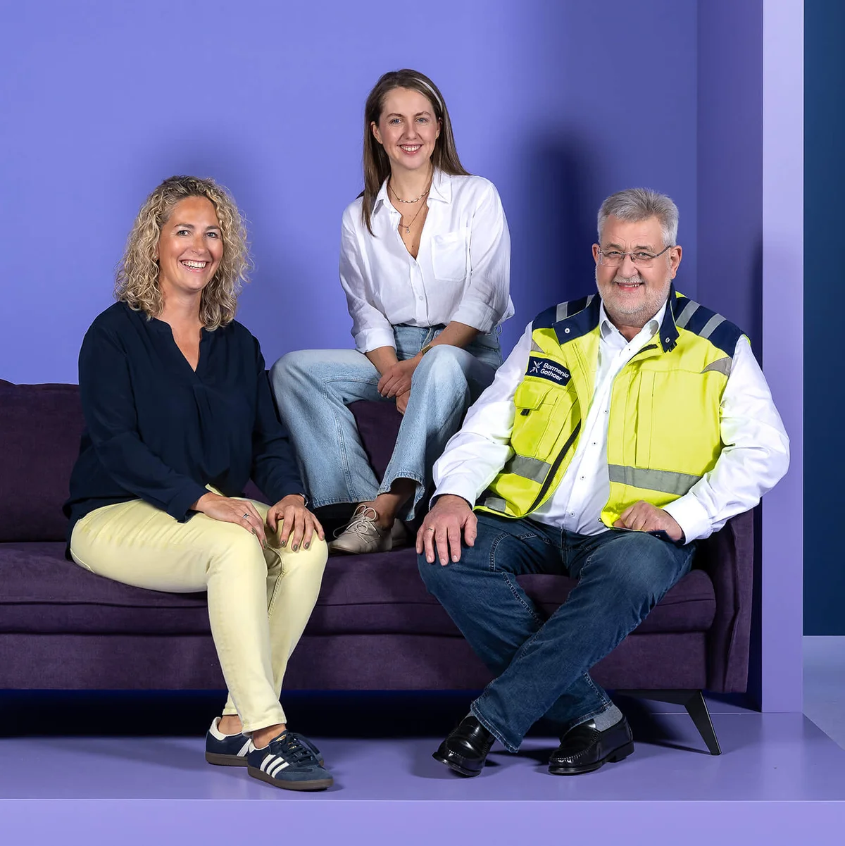 Drei Personen sitzen auf einem lila Sofa vor einem lila Hintergrund. Zwei Frauen tragen legere Kleidung, eine sitzt auf der Lehne. Ein Mann in einer gelben Sicherheitsweste sitzt rechts. Alle lächeln.