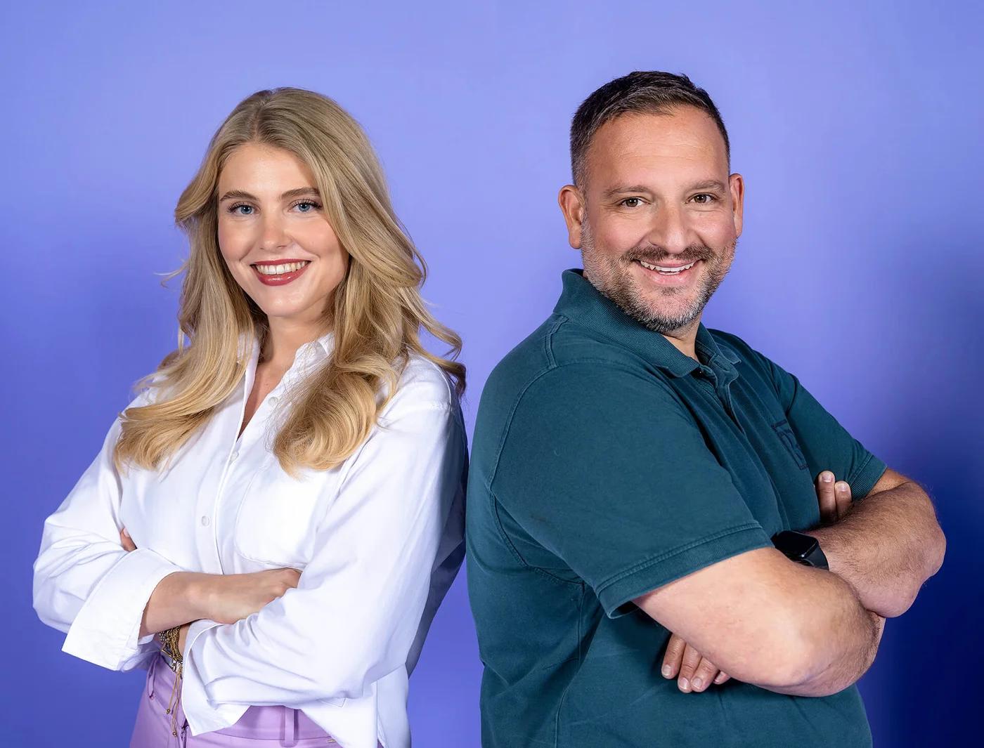 Zwei Personen stehen Rücken an Rücken vor einem lila Hintergrund. Die Frau links trägt eine weiße Bluse und der Mann rechts ein dunkelgrünes Hemd. Beide lächeln in die Kamera und haben die Arme vor der Brust verschränkt.