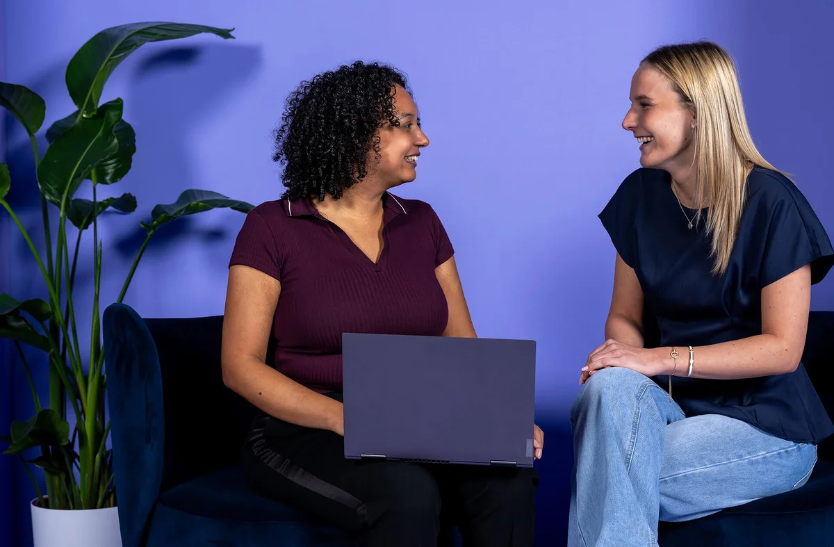 Zwei Frauen sitzen sich lächelnd gegenüber und unterhalten sich. Eine Frau hält einen Laptop auf dem Schoß. Die Szenerie ist entspannt und freundlich, vor einem lila Hintergrund, mit einer Pflanze im Hintergrund.