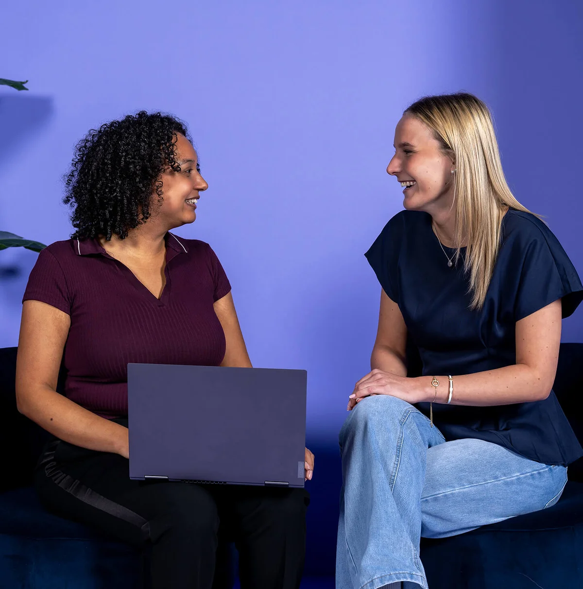 Zwei Frauen sitzen sich lächelnd gegenüber und unterhalten sich. Eine Frau hält einen Laptop auf dem Schoß. Die Szenerie ist entspannt und freundlich, vor einem lila Hintergrund, mit einer Pflanze im Hintergrund.