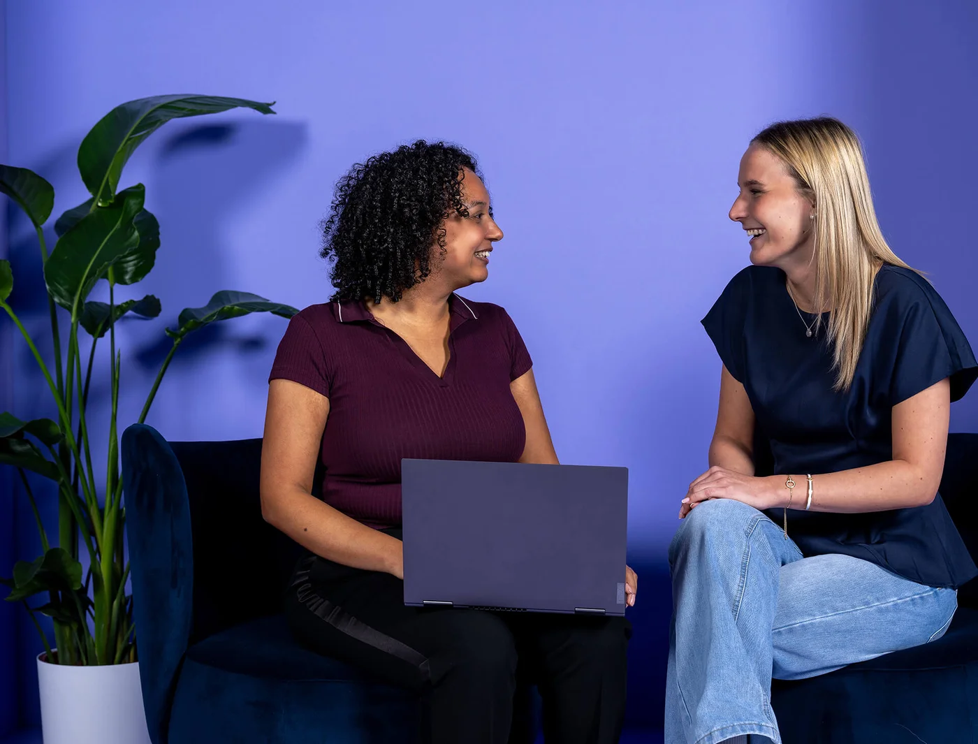 Zwei Frauen sitzen sich lächelnd gegenüber und unterhalten sich. Eine Frau hält einen Laptop auf dem Schoß. Die Szenerie ist entspannt und freundlich, vor einem lila Hintergrund, mit einer Pflanze im Hintergrund.